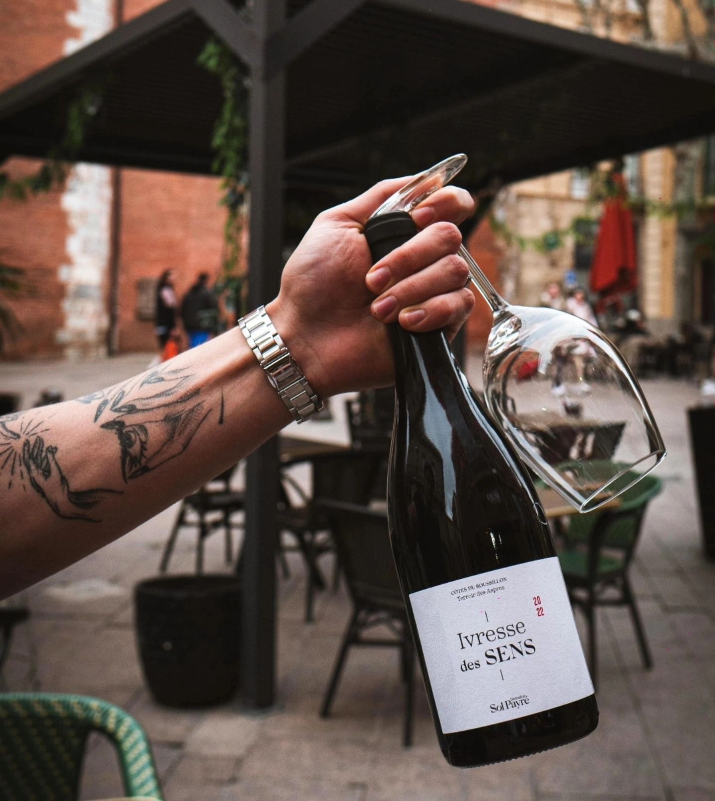 Personne tenant une bouteille de vin et un verre en extérieur ; tatouage visible, montre.