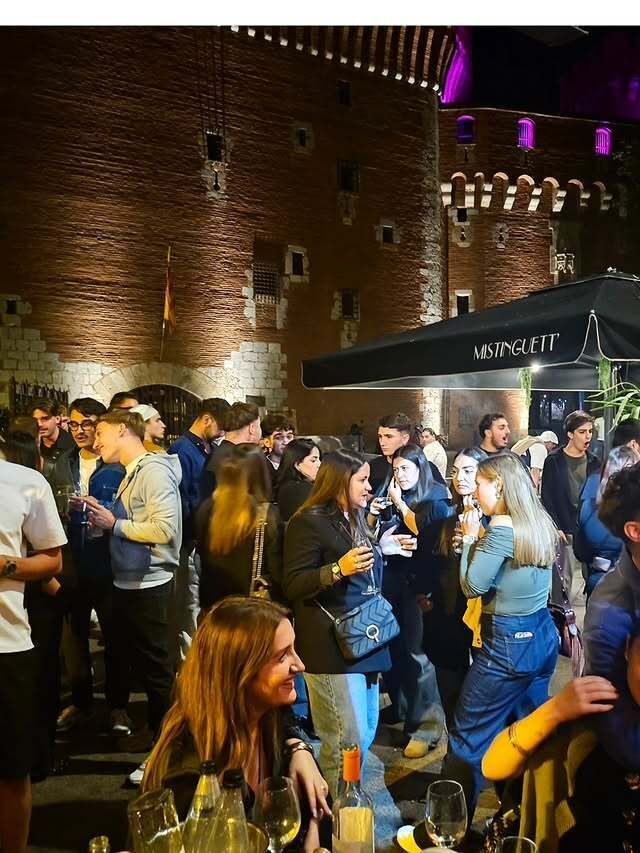 Des gens se rassemblent devant un bar, scène nocturne. Bâtiment en briques rouges en arrière-plan, parapluies, lumières.