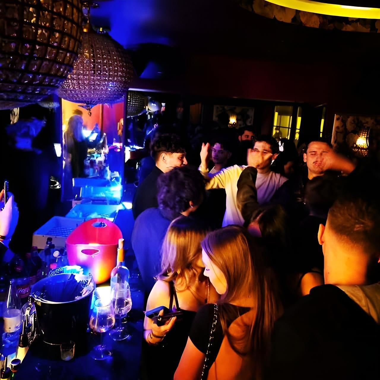 Dans un bar à l'éclairage tamisé, des gens se rassemblent près du comptoir et dansent.