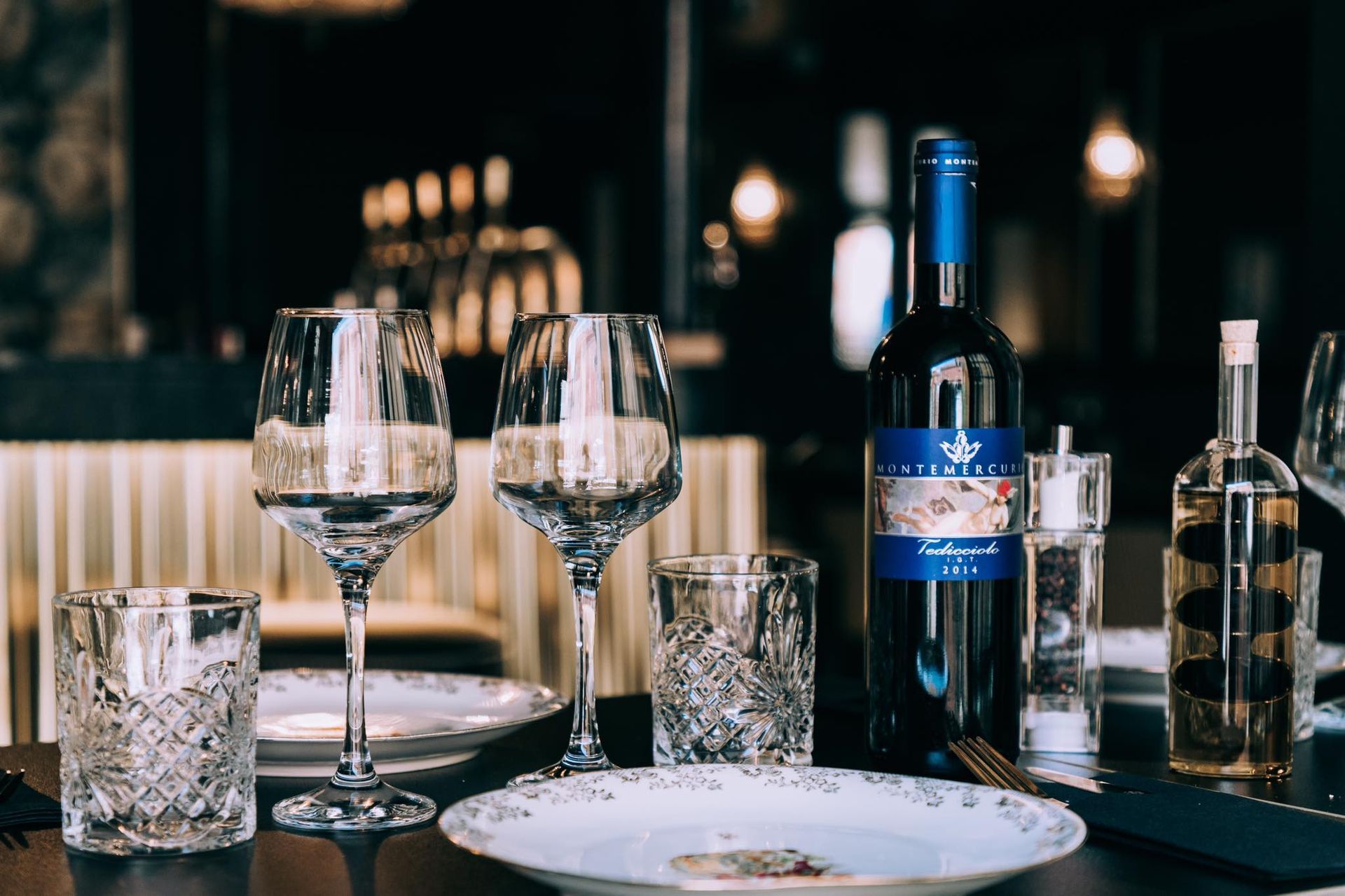 Dressage de table avec verres à vin, bouteille, assiettes et petits verres décoratifs, en intérieur, lumière tamisée.