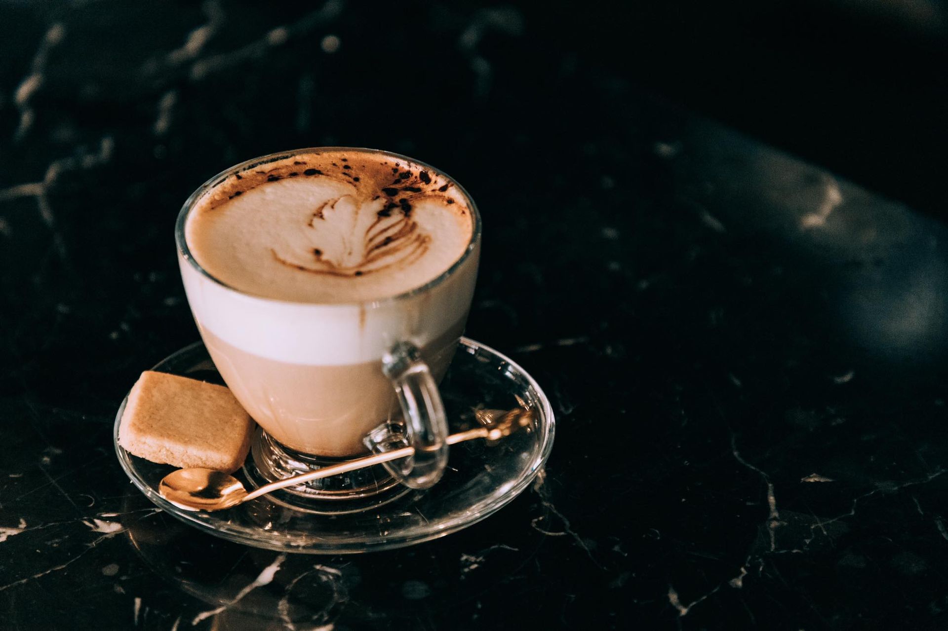 Du café dans une tasse en verre transparent, avec sa soucoupe et sa cuillère, posé sur une surface en marbre foncé. Un biscuit carré est disposé à proximité.