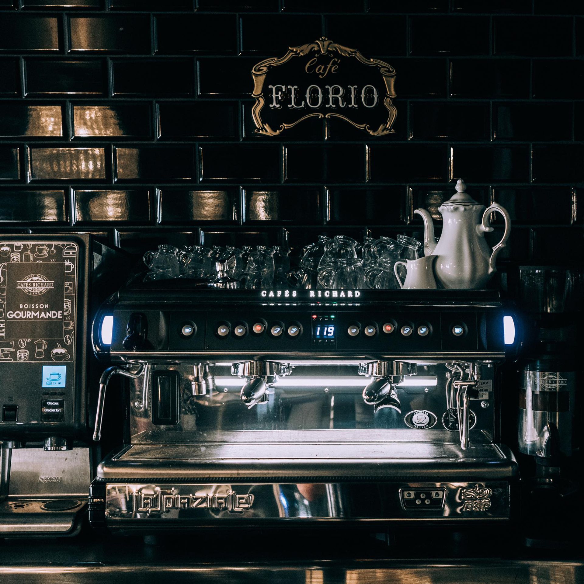 Machine à expresso, avec tasses en verre, théière blanche et fond de carrelage noir.