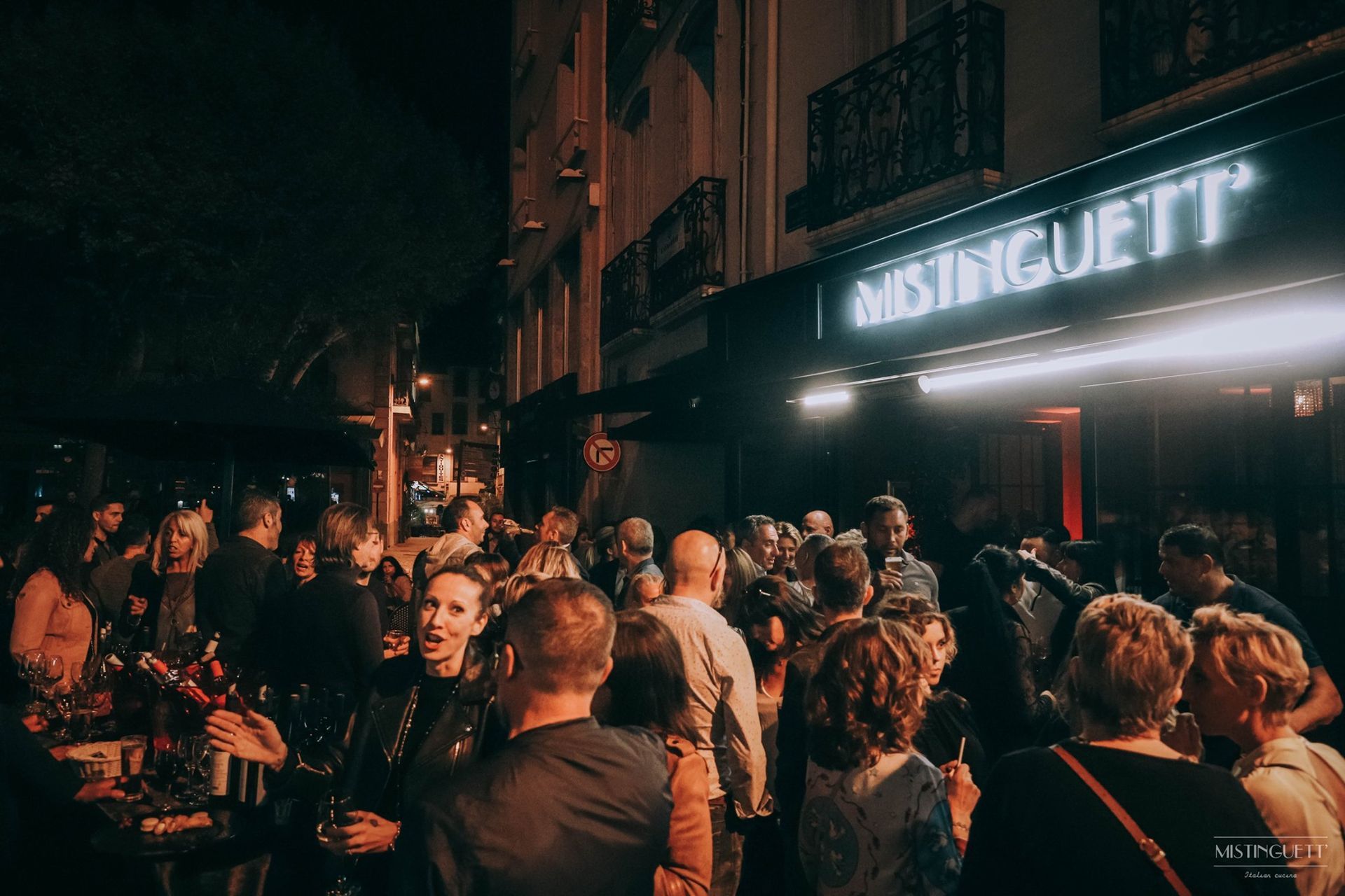 Des gens se rassemblent devant le restaurant, la nuit. Des lumières illuminent l'entrée.