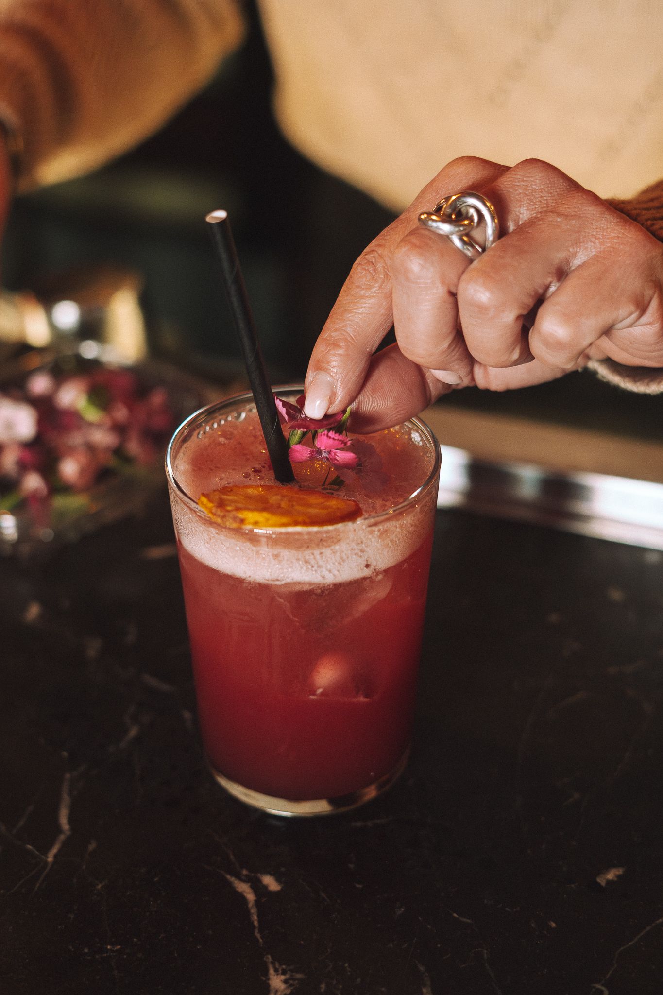 Une personne ajoute une petite fleur rose en garniture à un cocktail rouge avec une paille et une tranche d'orange, sur un comptoir noir.