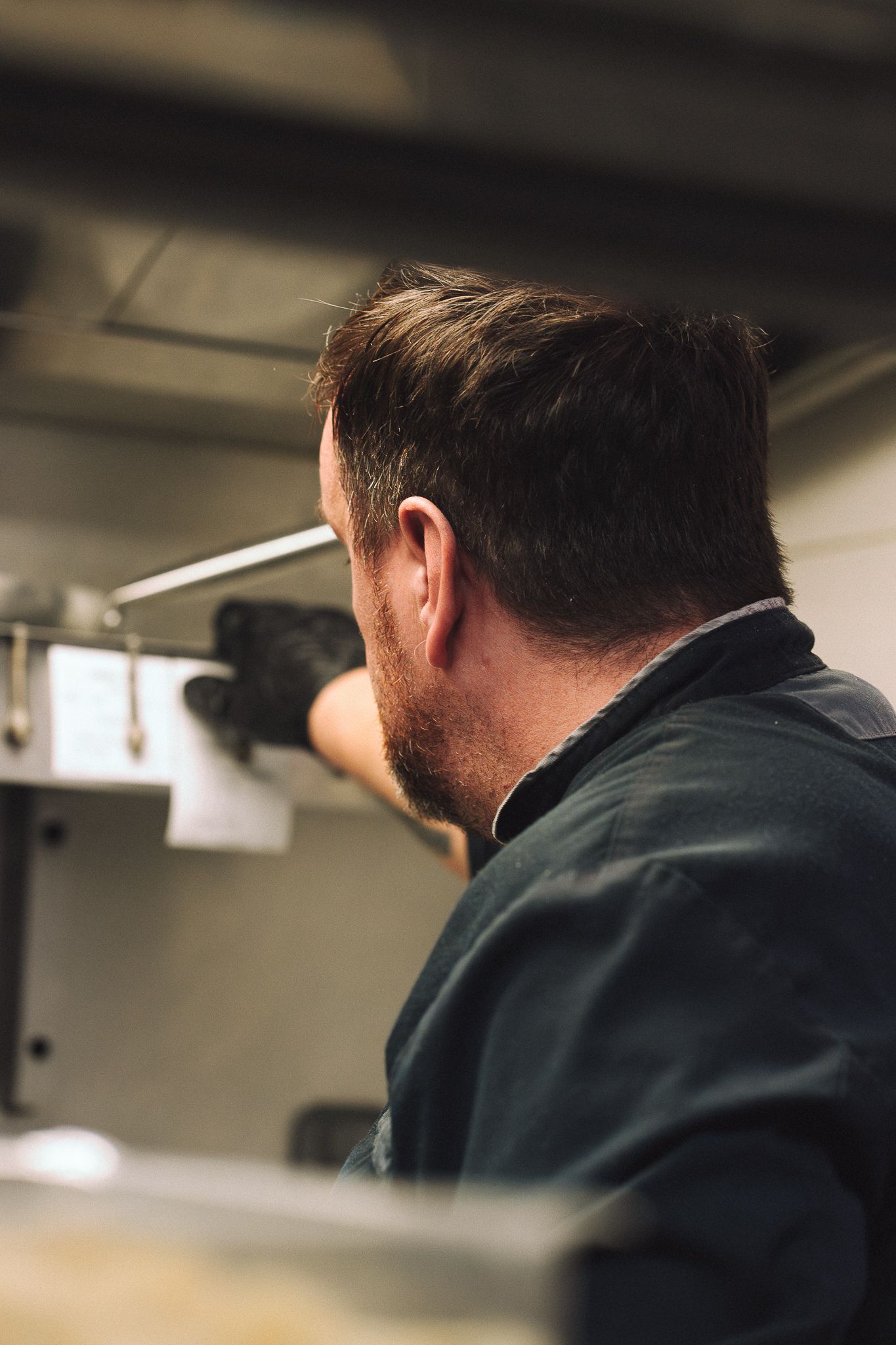 Un homme vêtu de sombre et portant des gants travaille sur une machine, à l'intérieur d'une cuisine professionnelle.
