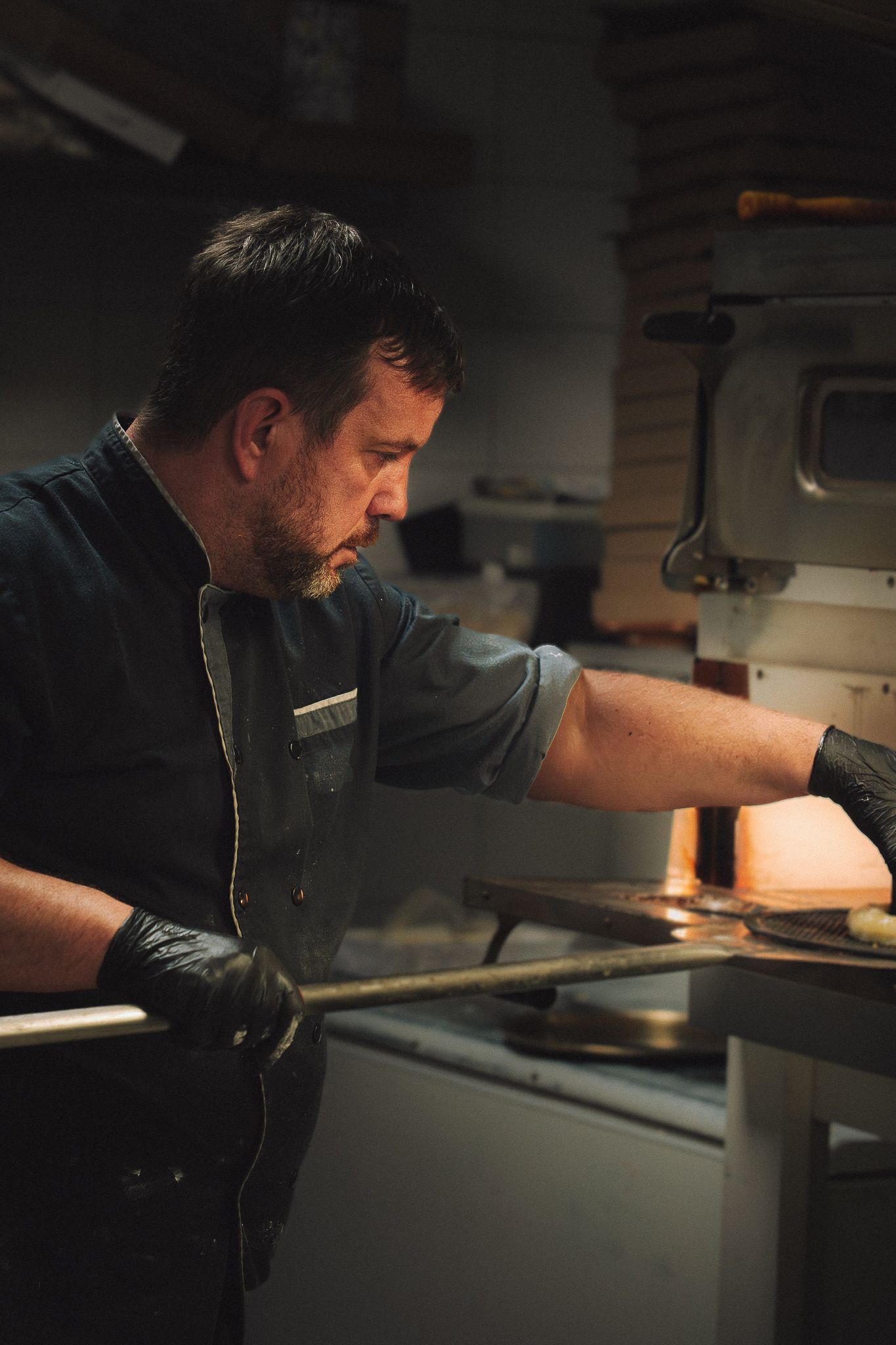 Un chef en uniforme sombre se trouve près d'un four à pizza, utilisant une pelle à pizza. Ambiance de cuisine tamisée.