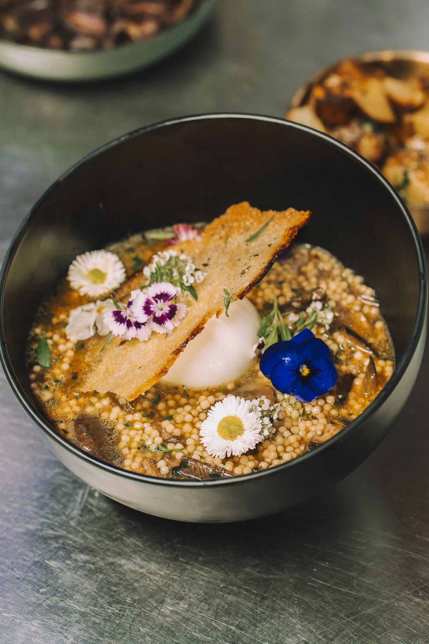 Bol de couscous garni de fleurs, de pain croustillant et de sauce blanche, présenté sur une surface réfléchissante.