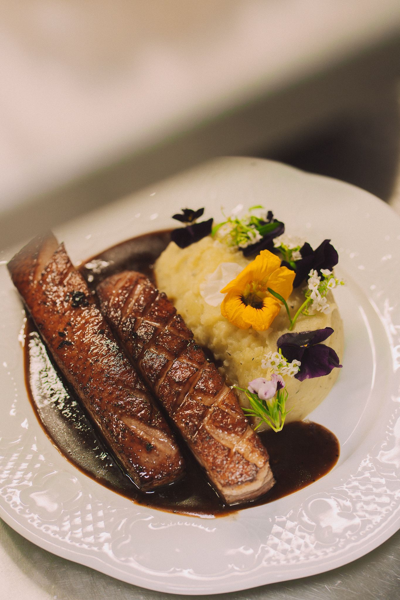 Magret de canard poêlé, sauce, purée de pommes de terre et fleurs comestibles sur une assiette blanche.