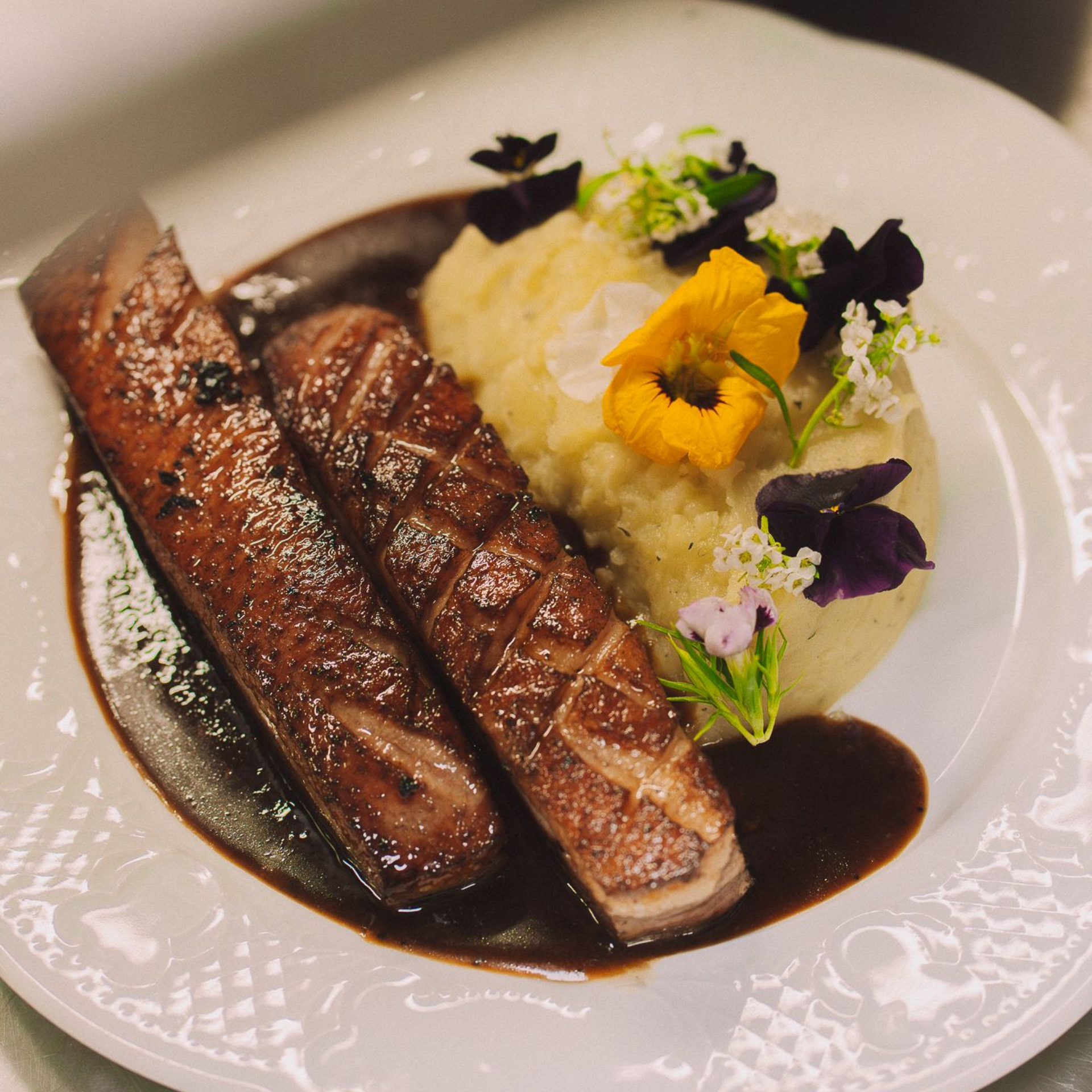 Magrets accompagnés de purée de pommes de terre, de sauce brune et de fleurs comestibles sur une assiette blanche.