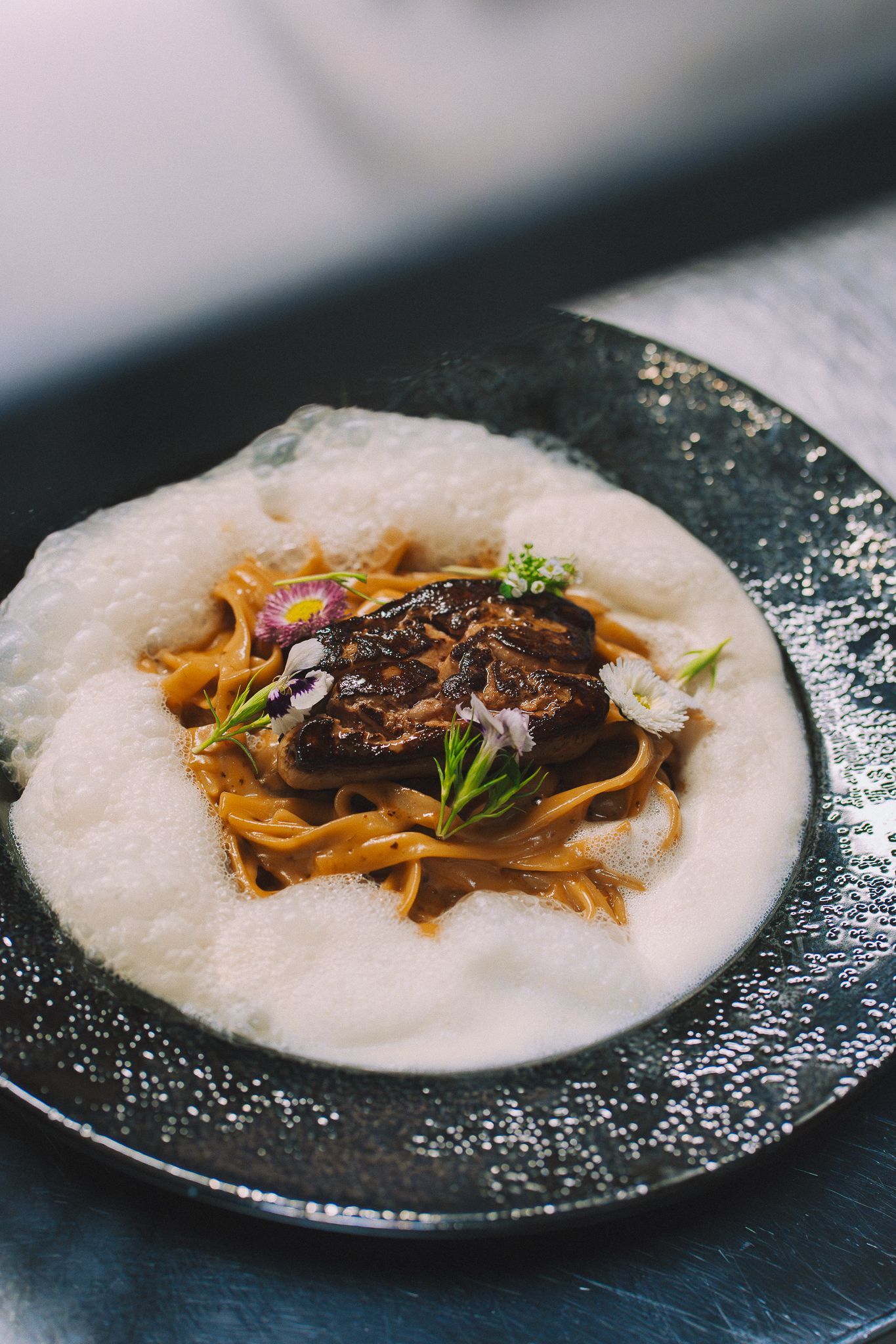 Plat de pâtes au foie gras poêlé, entouré d'une mousse, sur une assiette sombre.