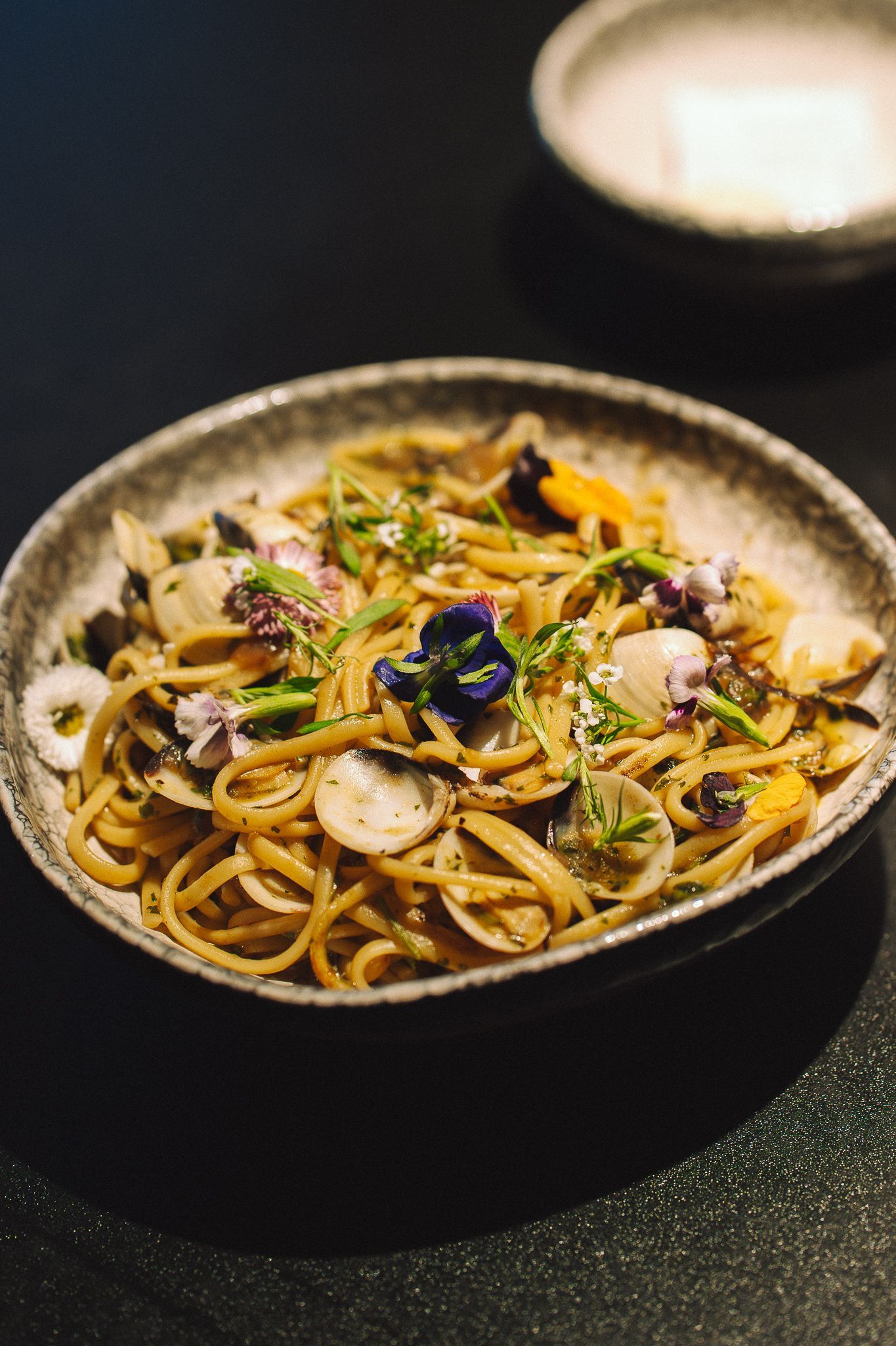 Plat de pâtes dans un bol gris moucheté, garni de fleurs et d'herbes sur un fond sombre.
