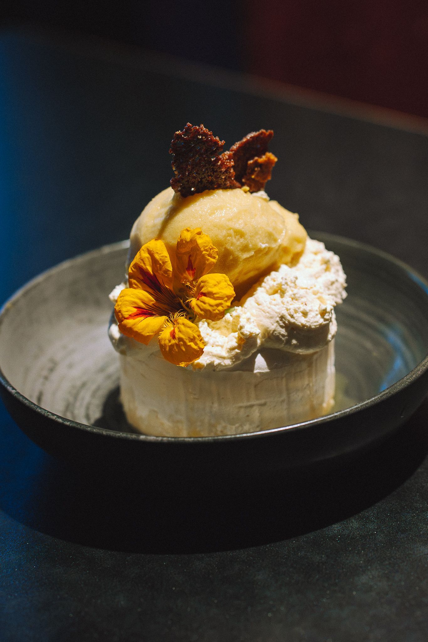 Dessert composé de glace, de fleurs comestibles et de garniture, présenté dans un bol sombre sur une surface noire.
