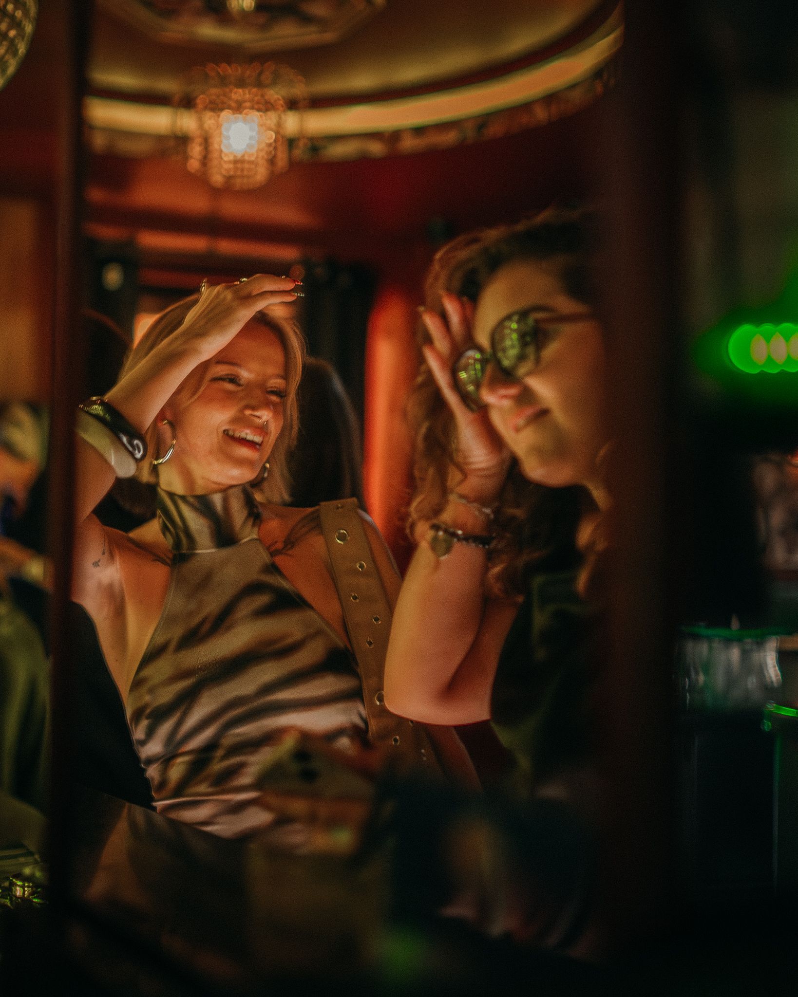 Une personne vêtue d'un haut à imprimé zèbre prend un selfie devant un miroir dans un bar à l'éclairage tamisé et aux tons chauds, avec un ami visible dans le reflet.