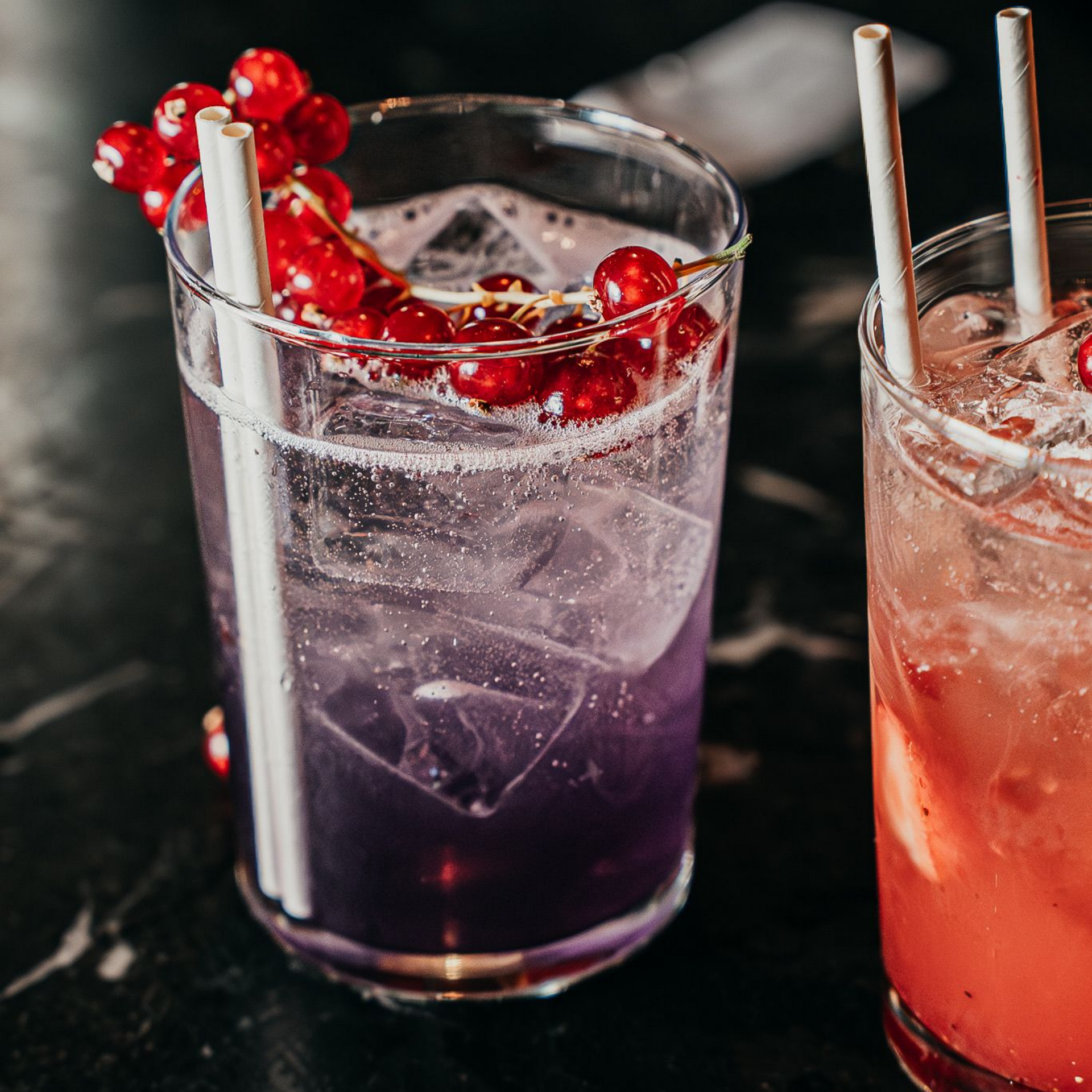 Cocktail servi dans un verre avec un liquide violet, des glaçons et des baies rouges, garni de pailles en papier.