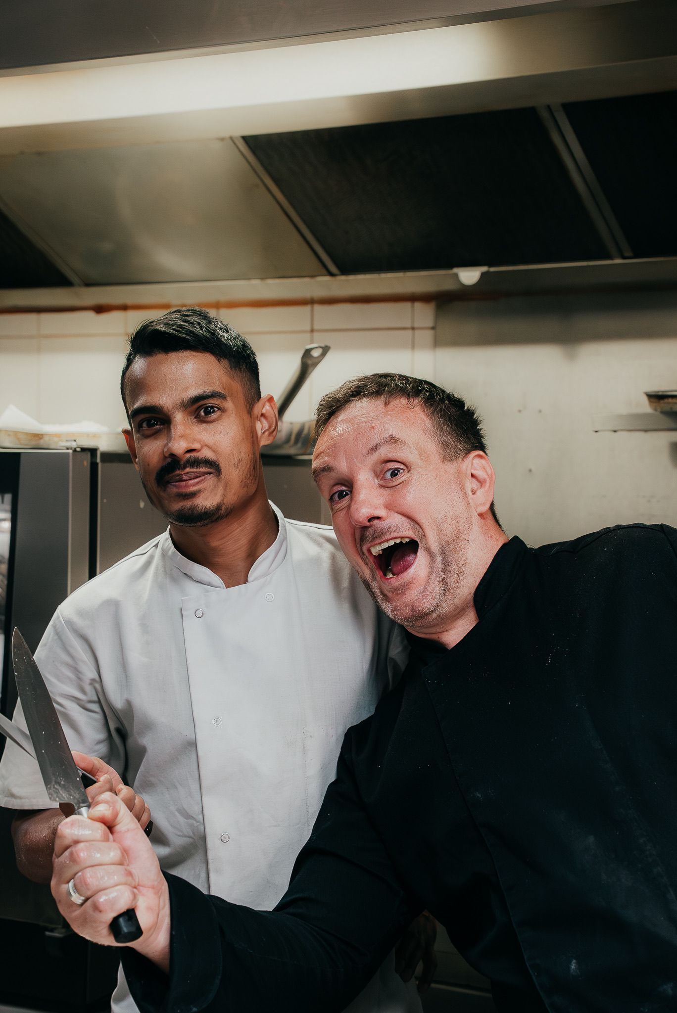 Deux chefs dans une cuisine ; l’un tient un couteau et sourit, l’autre prend la pose. Éclairage fluorescent vif.