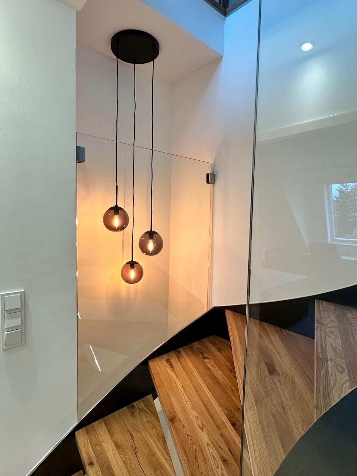 Eine hölzerne Wendeltreppe mit einem Glasgeländer und von der Decke hängenden Lichtern.