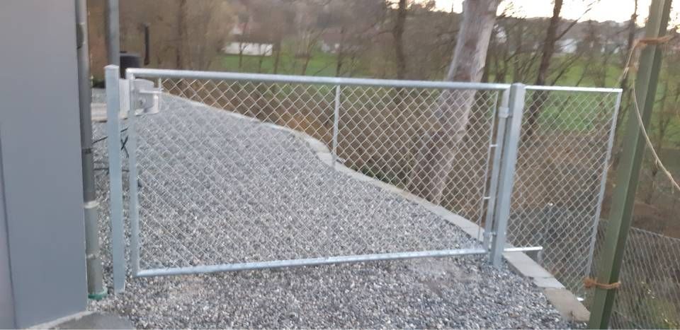 Ein geschlossenes Maschendrahttor versperrt eine Kiesauffahrt neben einem Gebäude und einem Waldstück.