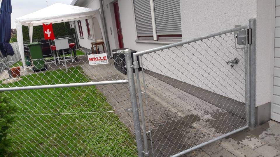 Ein Maschendrahttor neben einem weissen Zelt in einem Hinterhof, im Hintergrund eine rote Schweizer Flagge.
