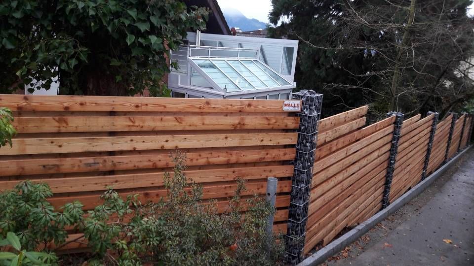 Holzzaun am Strassenrand, dahinter eine Steinsäule und Grünflächen