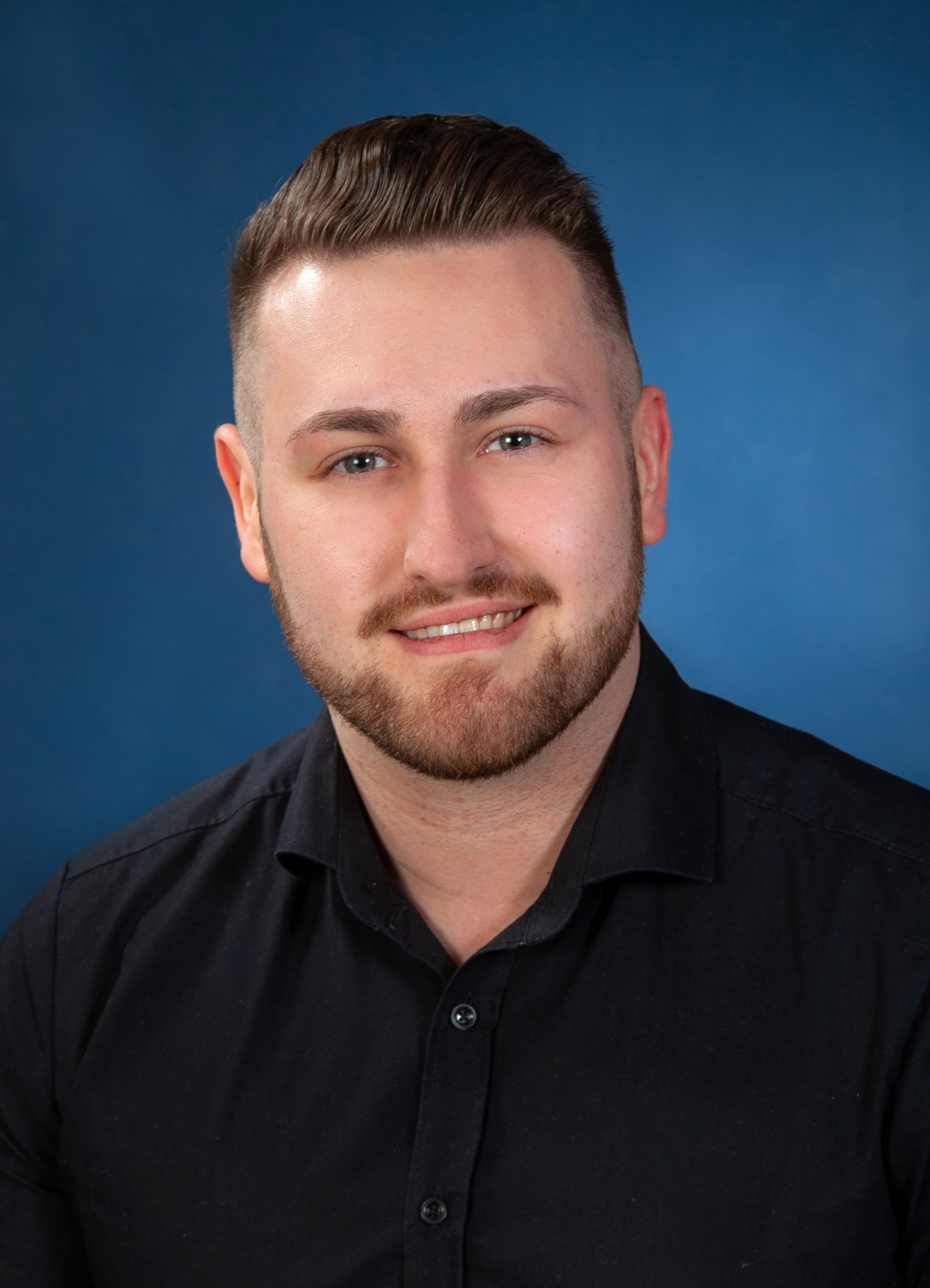 Ein Mann mit Bart trägt ein schwarzes Hemd und lächelt in die Kamera.