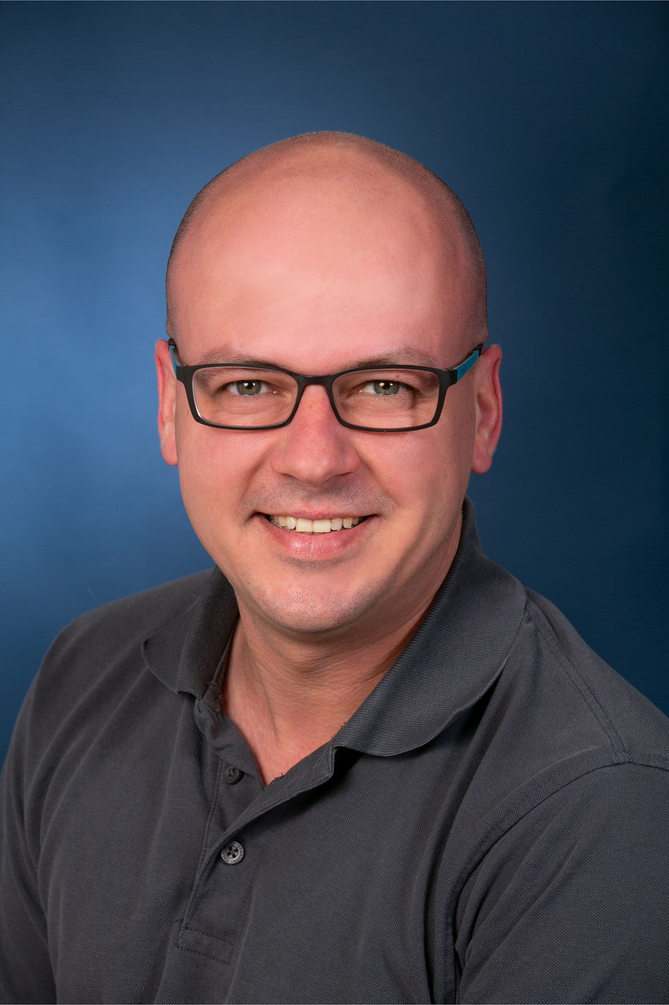 Ein kahlköpfiger Mann mit Brille und grauem Hemd lächelt in die Kamera.