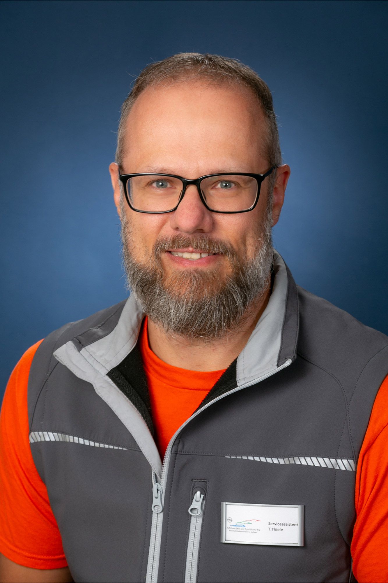 Ein Mann mit Bart und Brille trägt eine Weste und ein orangefarbenes Hemd.