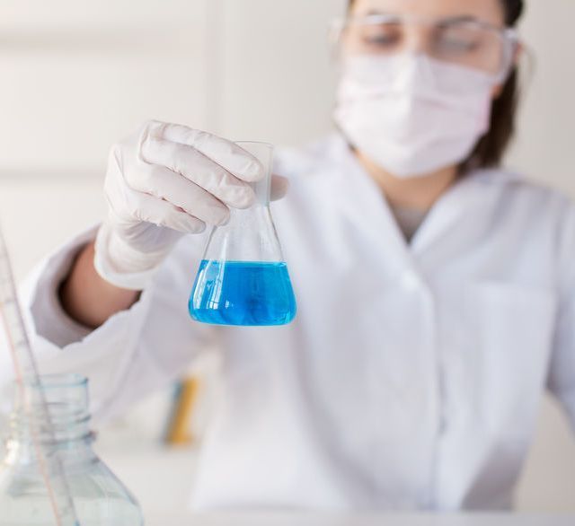 Un científico que lleva una máscara y guantes sostiene un vaso de precipitados con líquido azul.