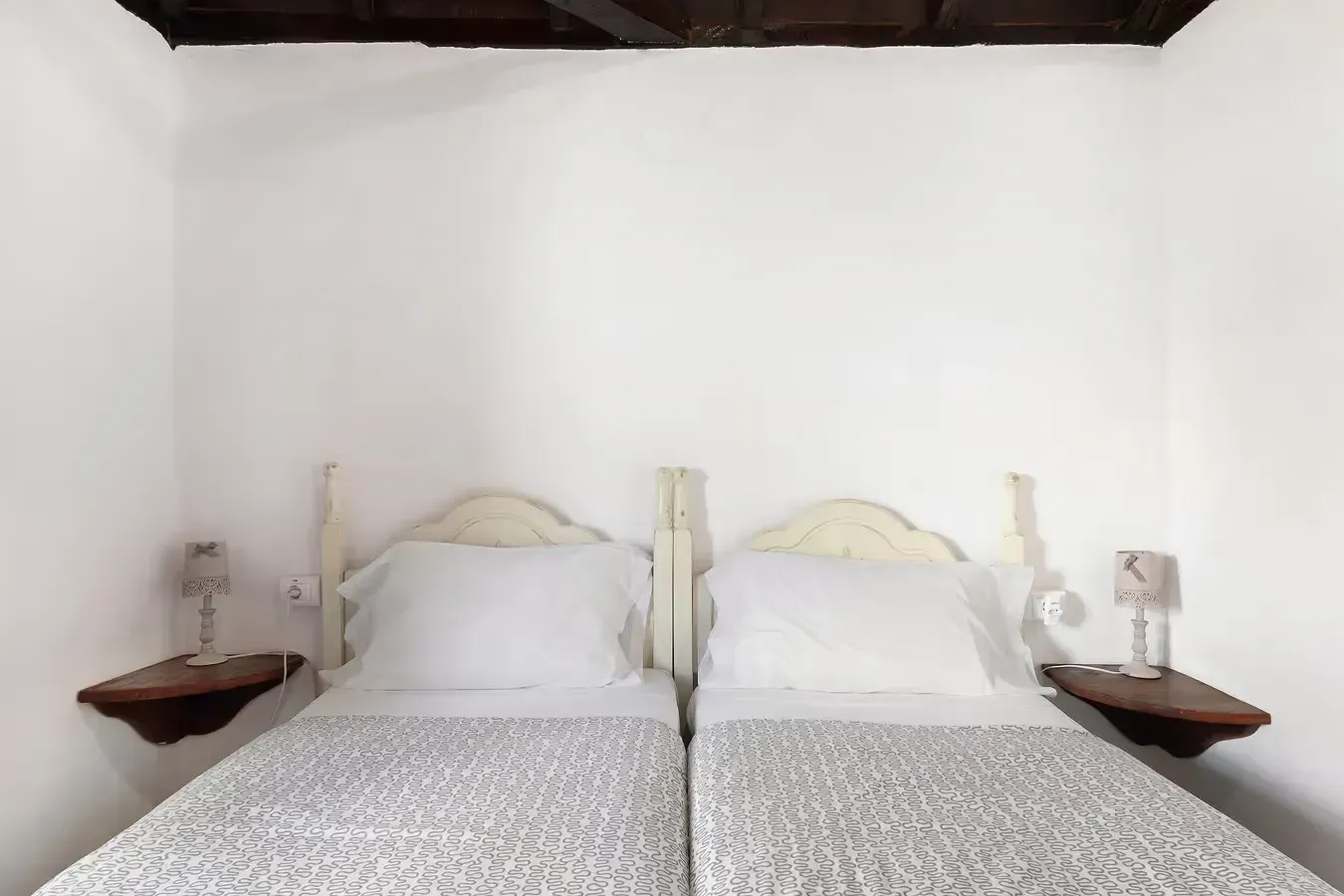 Dos camas individuales con cabeceros blancos, mesitas de noche y lámparas contra una pared blanca.