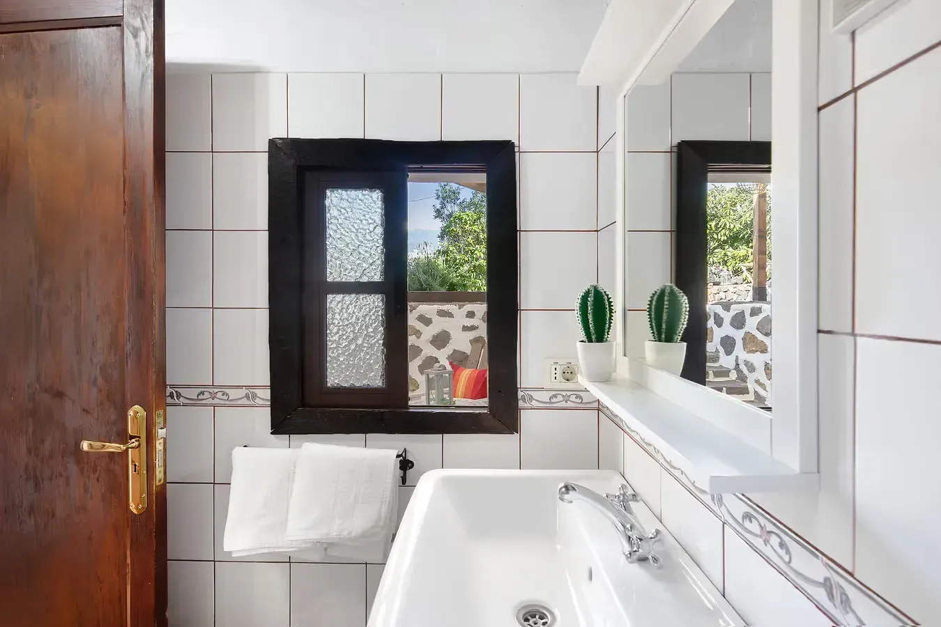 Baño con paredes de azulejos blancos, una ventana de marco oscuro y un lavabo blanco.