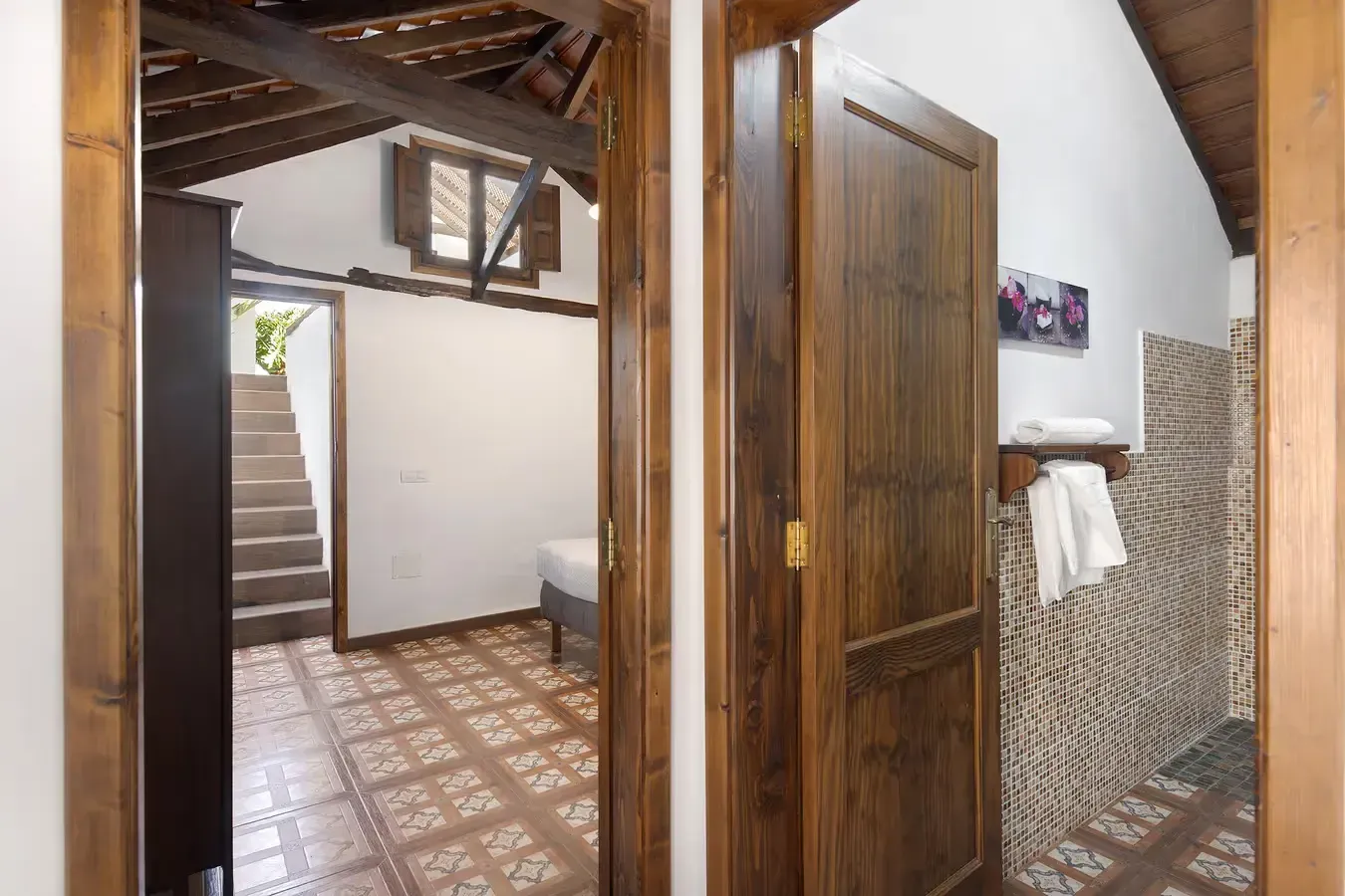 Puertas de madera que dan a un dormitorio y un baño. Suelo de baldosas estampadas y vigas vistas en el techo.