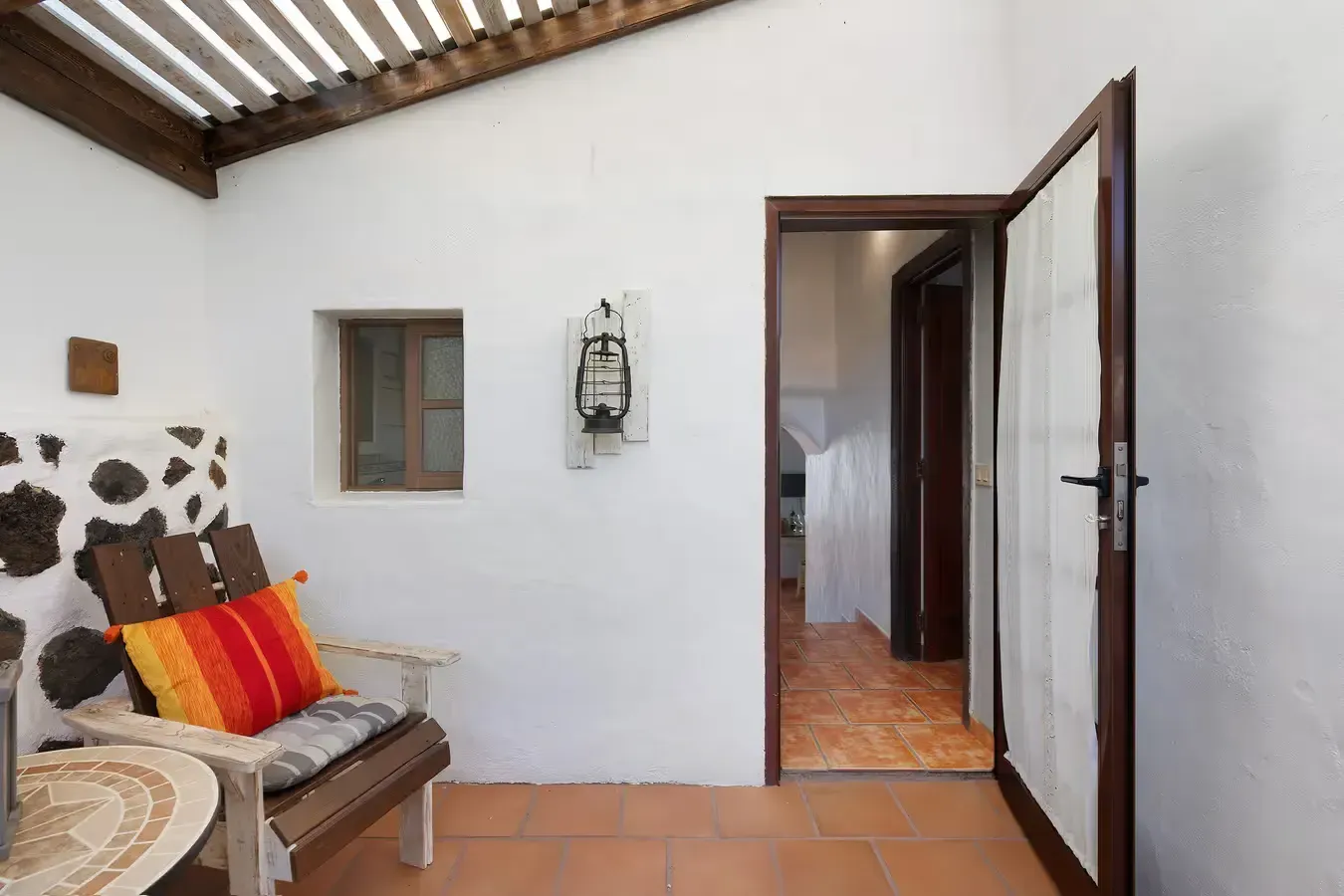 Patio cubierto con banco, pequeña ventana y puerta abierta que da a un pasillo.