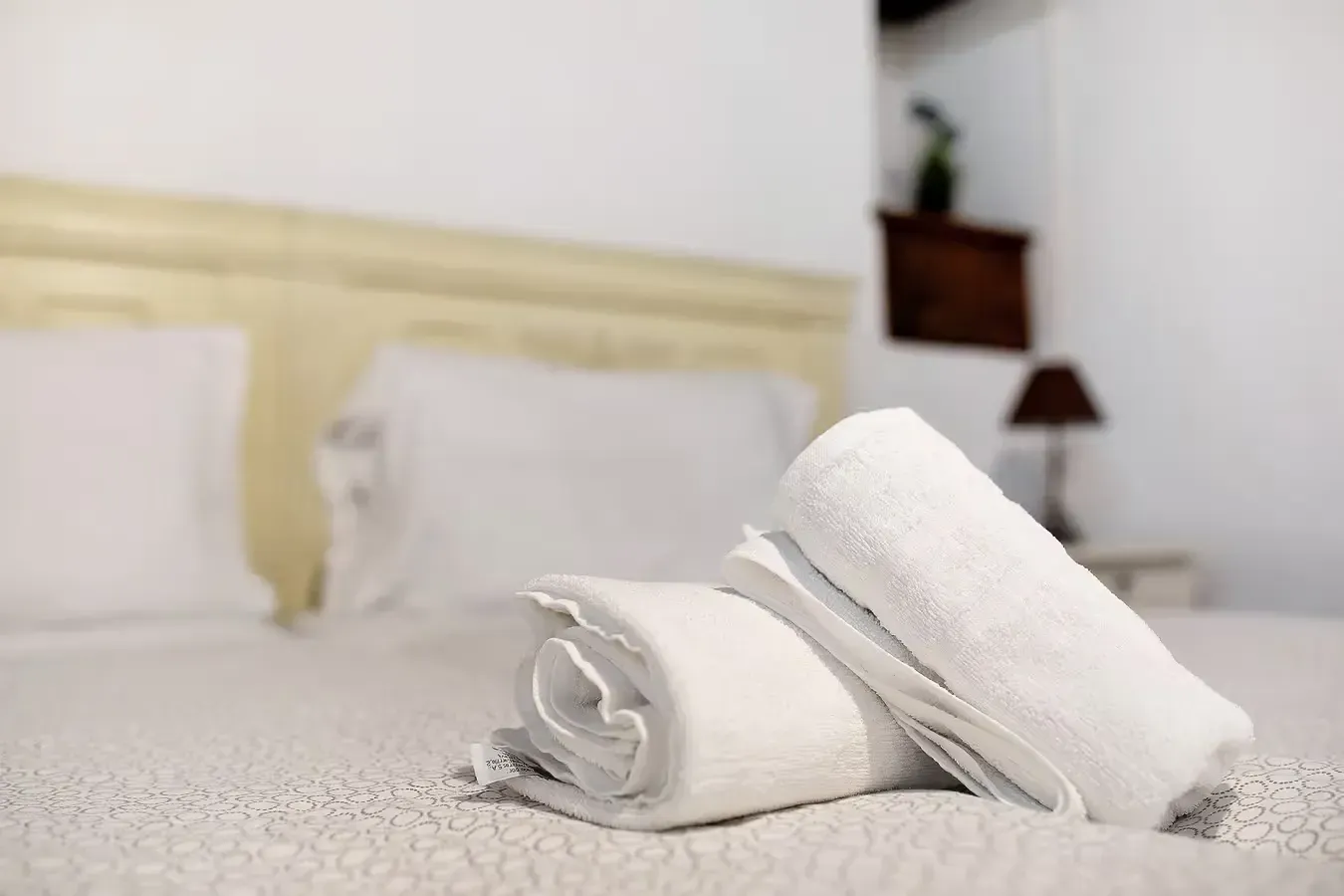 Toallas blancas enrolladas sobre una cama con ropa de cama blanca, cabecero en una habitación luminosa.