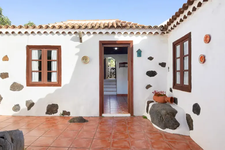 Patio de una casa de estuco blanco con ventanas y puerta marrones, piso de baldosas de terracota y cielo azul.