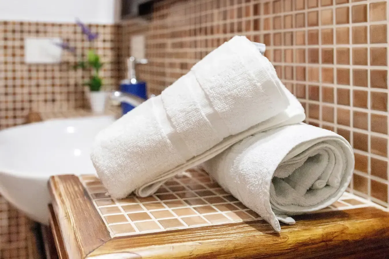 Dos toallas blancas enrolladas sobre una superficie de baldosas en un baño, cerca de un lavabo.