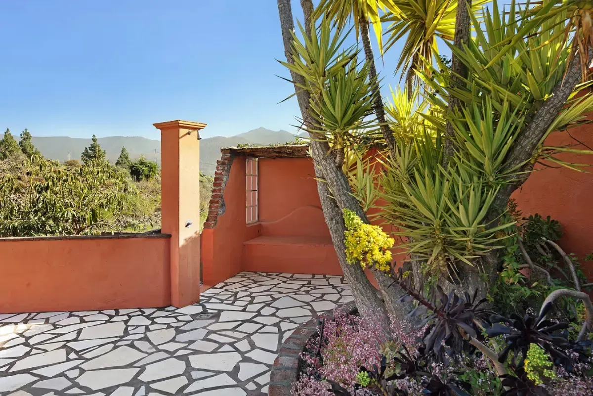 Terraza con baldosas de piedra, paredes naranjas y un árbol con follaje verde, con vistas a las montañas.