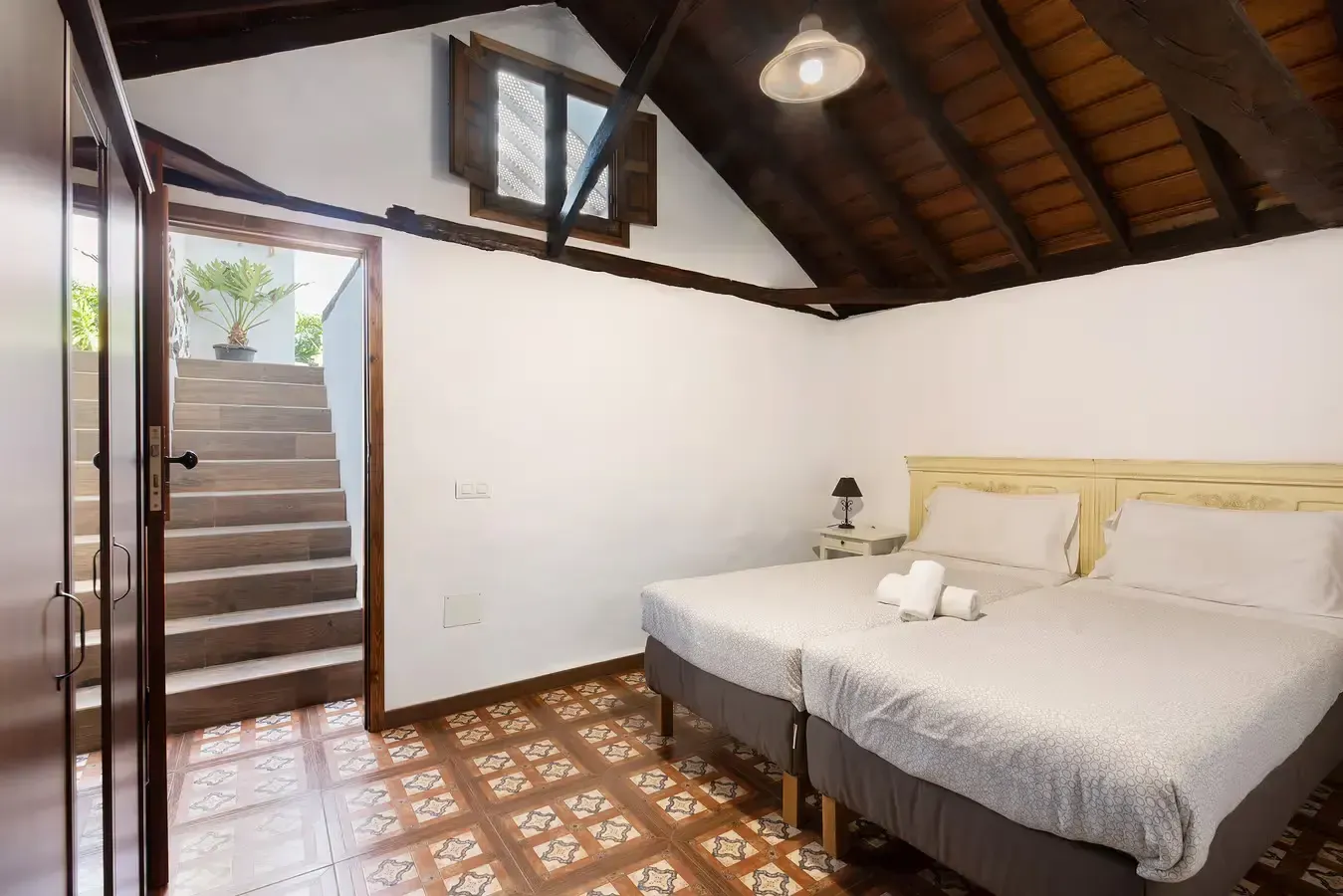 Dormitorio con dos camas individuales, suelo de baldosas, techo de madera y escalera a la vista.