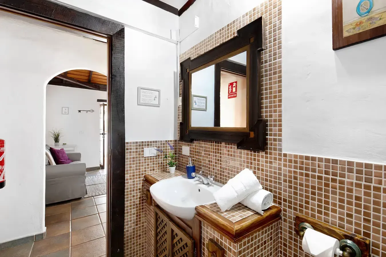 Baño con paredes de azulejos marrones, lavabo, espejo y puerta a una sala de estar.