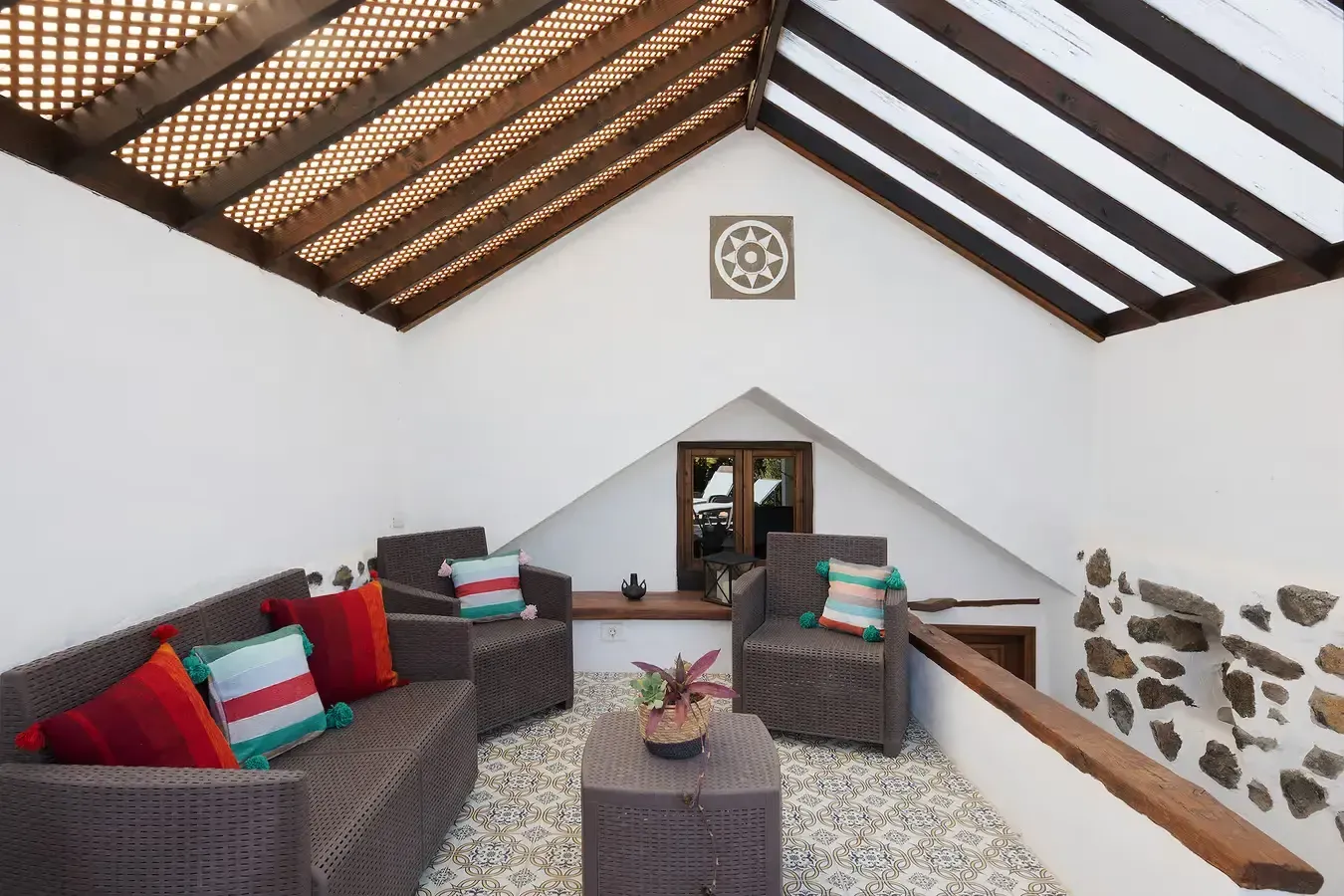 Zona de estar al aire libre con muebles de mimbre marrón, almohadas rojas y turquesas y suelo de piedra