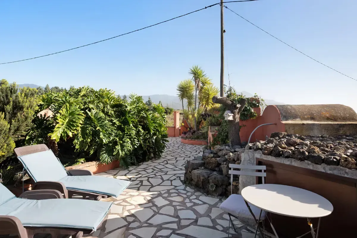 Patio con reposeras, mesita y camino de piedra, rodeado de vegetación y un cielo azul claro.