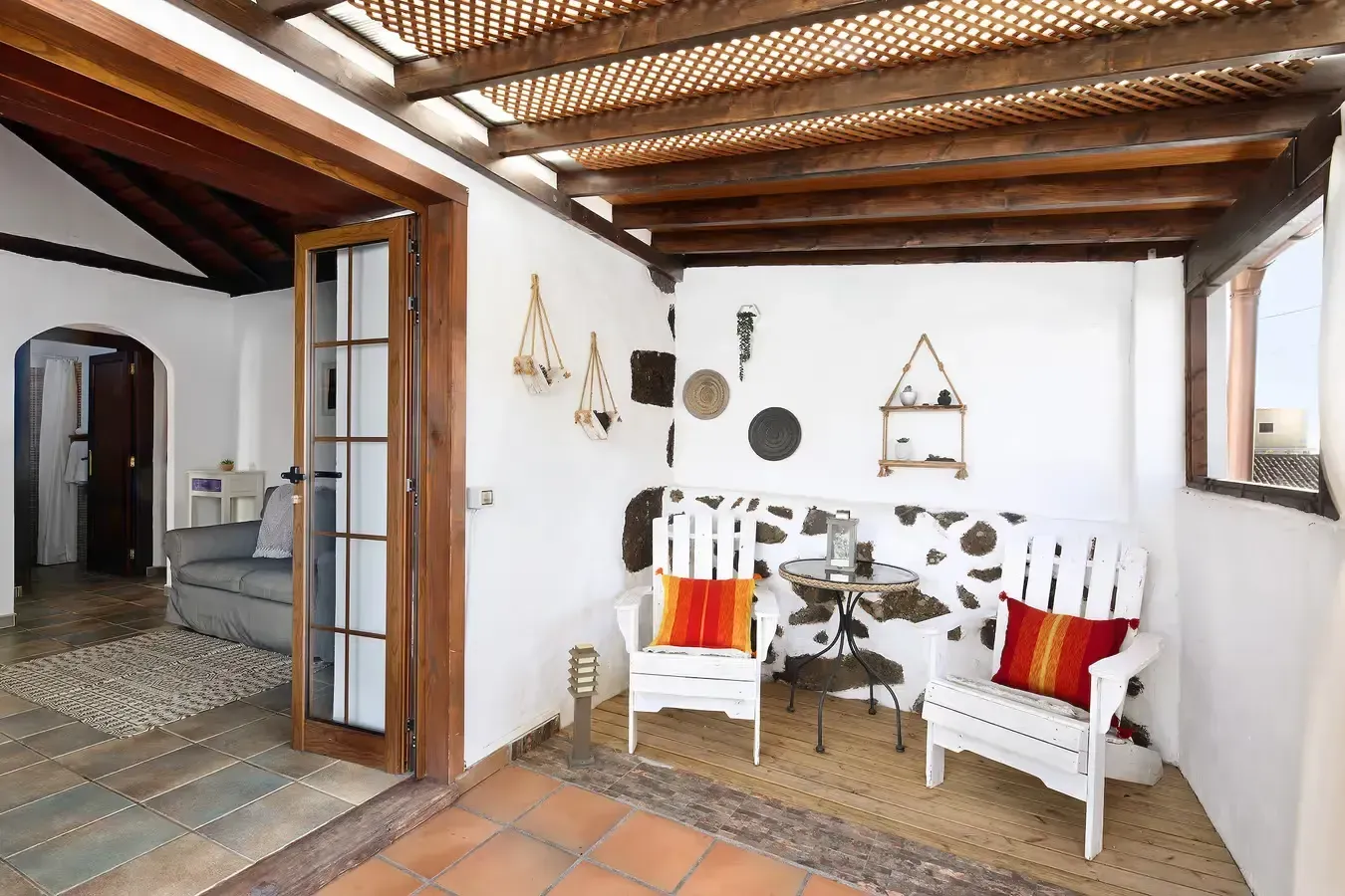 Patio cerrado con sillas blancas, elementos decorativos, techo de madera y puerta de entrada a un espacio habitable.