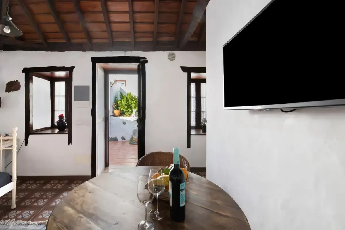 Habitación interior con paredes blancas, una mesa de madera con copas de vino y un televisor montado en la pared.