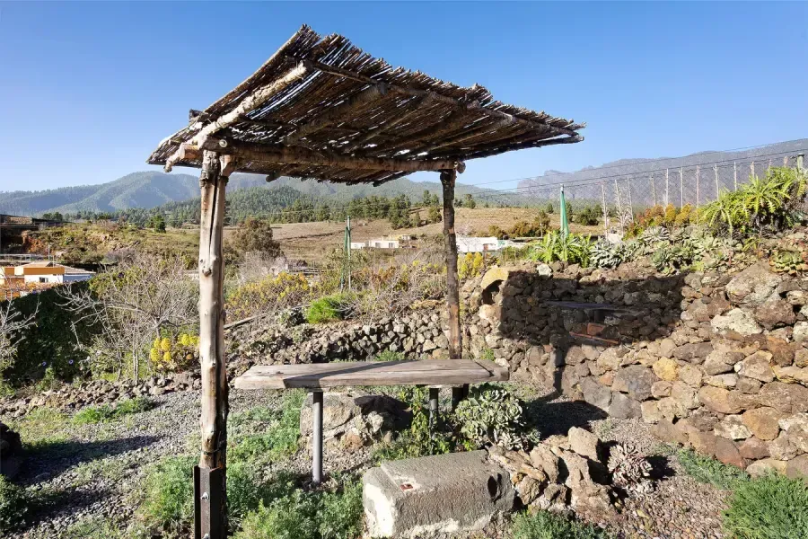 Refugio de madera con techo de paja, con vistas a un paisaje rural con montañas bajo un cielo azul.