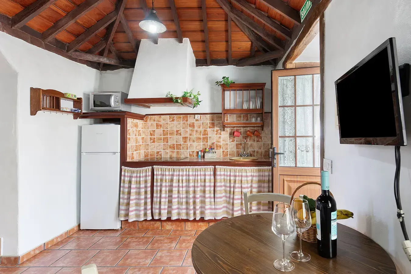 Cocina con salpicadero de azulejos, frigorífico, TV, mesa con vino y techo rústico de madera.