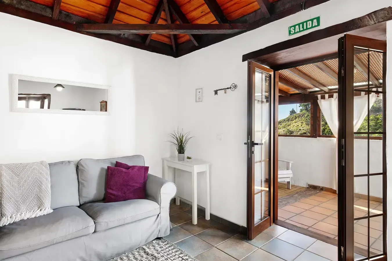 Interior de una habitación de paredes blancas con un sofá gris, que da a un balcón con vista.