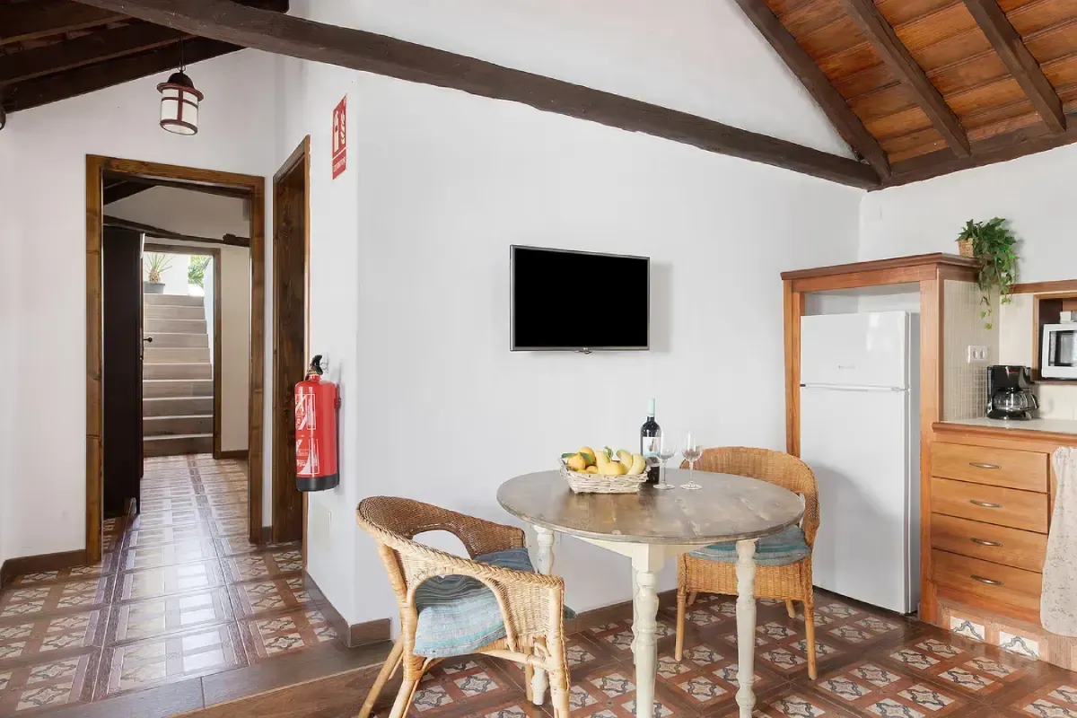 Interior de una habitación con TV, mesa, sillas, nevera y una puerta que da a unas escaleras.