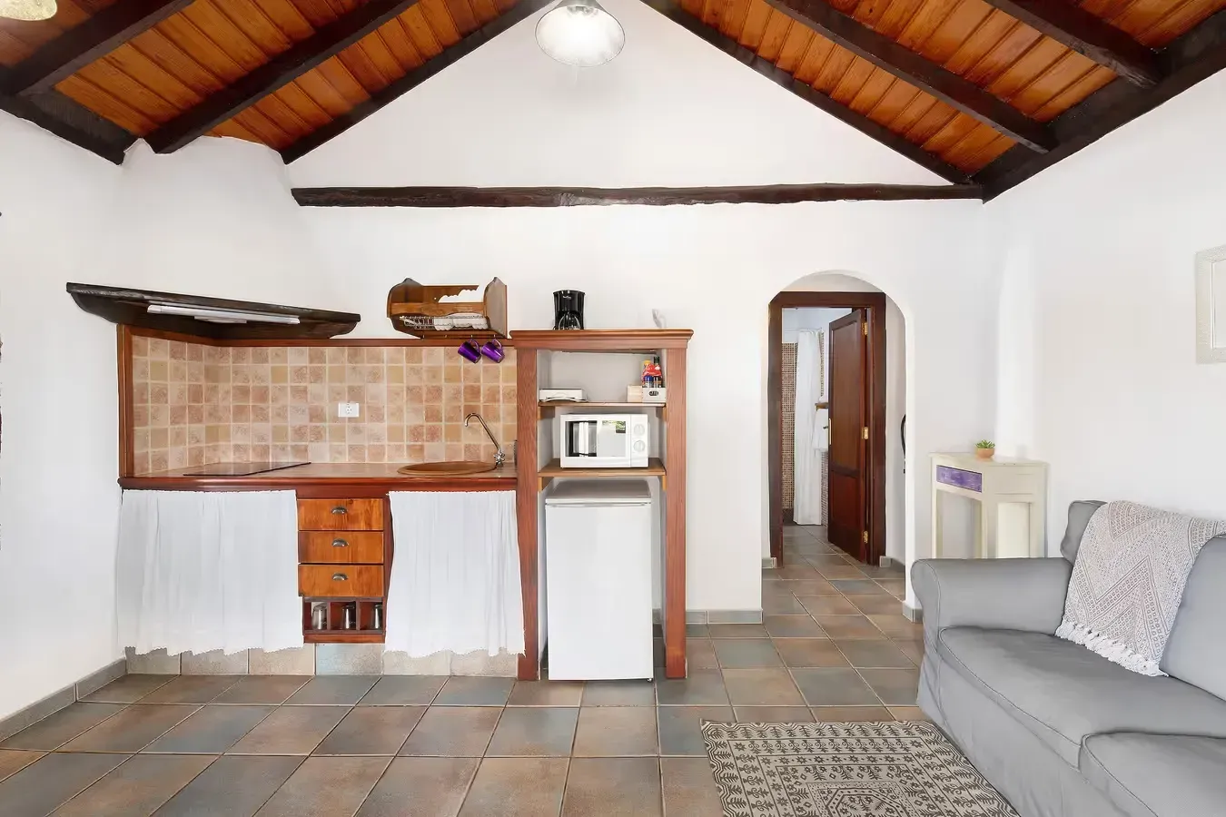 Interior de una cocina y sala de estar, con gabinetes de madera, un refrigerador pequeño, un sofá gris y una puerta abierta.
