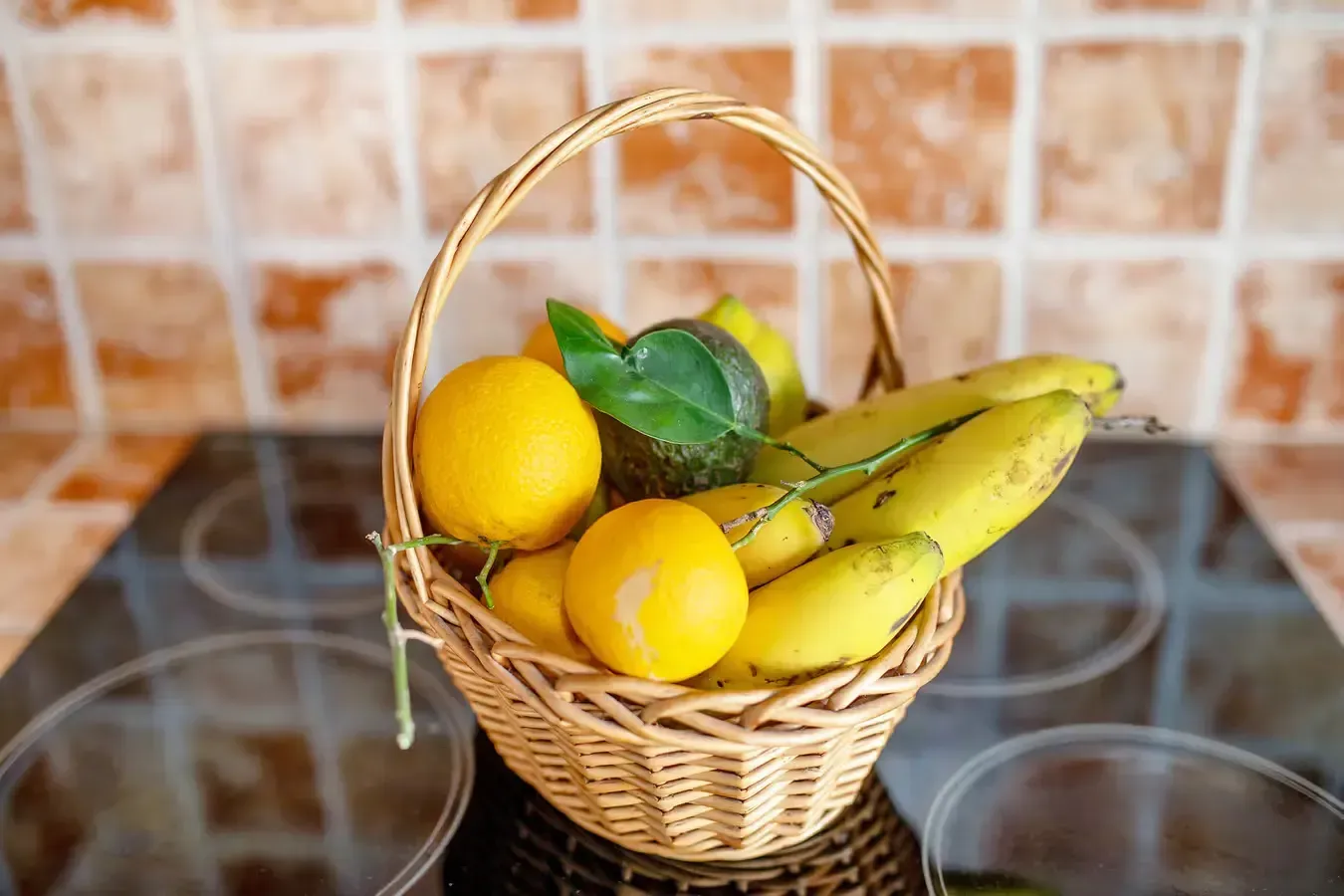Cesta de frutas: limones, plátanos y un aguacate sobre una estufa negra, contra una pared de azulejos.