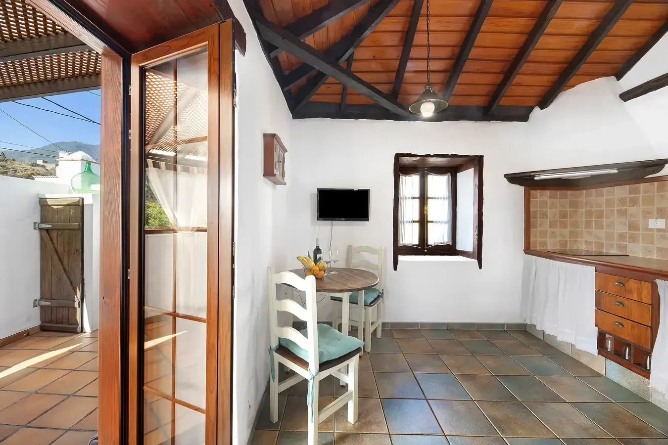 Interior con piso de terracota, paredes blancas, techo y ventana de madera, pequeña área de cocina