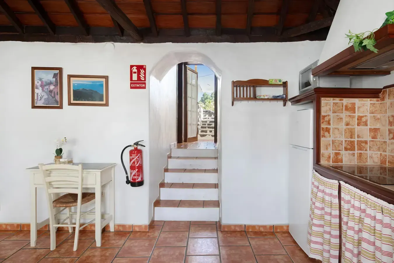 Interior de una cocina con puerta, escalones y una mesa blanca. Hay un extintor en la pared.