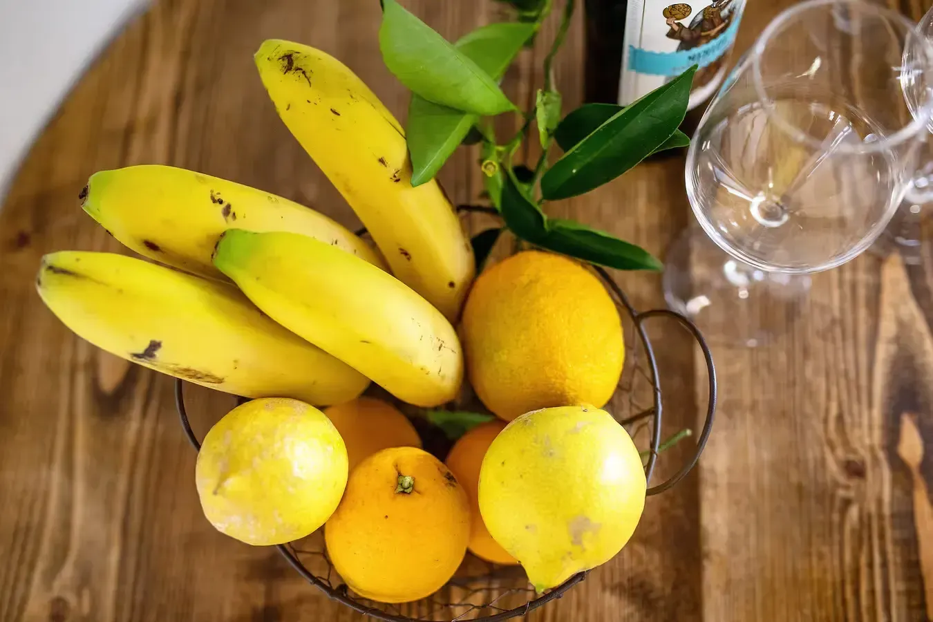 Cesta de plátanos amarillos, limones y naranjas sobre una mesa de madera con un vaso y una planta parcialmente visibles.